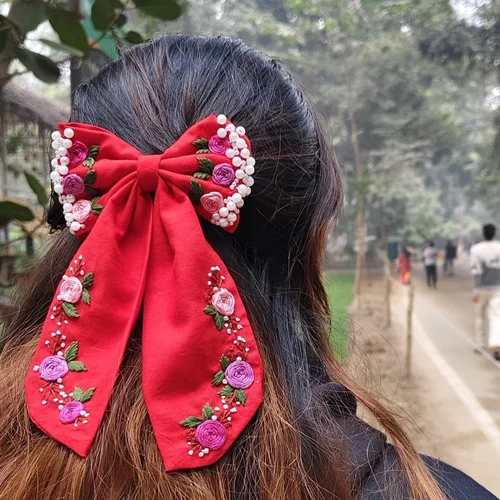 Red fabric hair bow with floral embroidery, pearl embellishments, and long ribbon tails